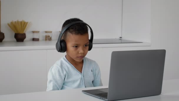 Онлайн-образование. Маленький черный мальчик в наушниках учится с ноутбуком дома — стоковое видео