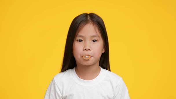Маленькая корейская девочка жует и пускает жвачку над жёлтым фоном — стоковое видео