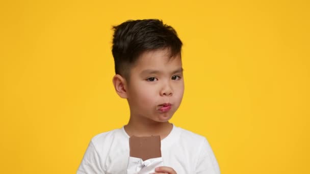 Pequeño chico japonés comiendo chocolate bar sobre amarillo estudio fondo — Vídeo de stock