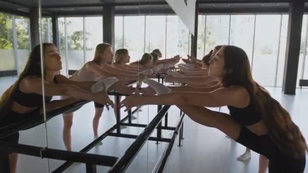 Clases de ballet. Zoom en tiro de bailarinas grupo de entrenamiento en estudio de baile, inclinándose hacia adelante con las piernas en la barra en el espejo — Vídeos de Stock