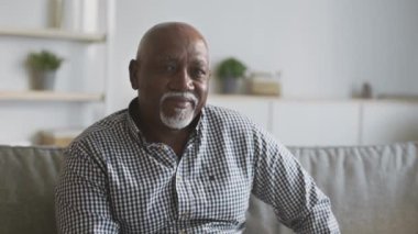 Mutlu yaşlanma. Pozitif kıdemli Afro-Amerikalı beyefendi evinde koltukta oturuyor ve kameraya gülümsüyor.