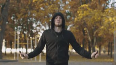 Yoğun kardiyo eğitimi ve vücut kurulanması. Kapüşonlu genç adam sonbahar parkında ip atlama alıştırması yapıyor.