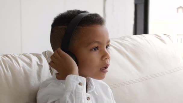 Malý černý chlapec nosit bezdrátové sluchátka poslech hudby zatímco sedí na gauči — Stock video