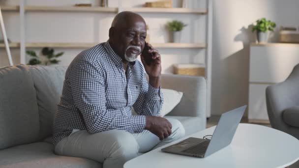 Freiberufler und Rentner. Senior afrikanisch-amerikanischer Mann berät Kunden beim Telefonieren und Arbeiten am Laptop — Stockvideo