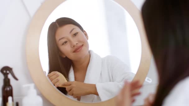 Haarpflegekonzept. Junge fröhliche asiatische Frau bürstet sich die Haare, schaut in den Spiegel im Badezimmer und lächelt — Stockvideo