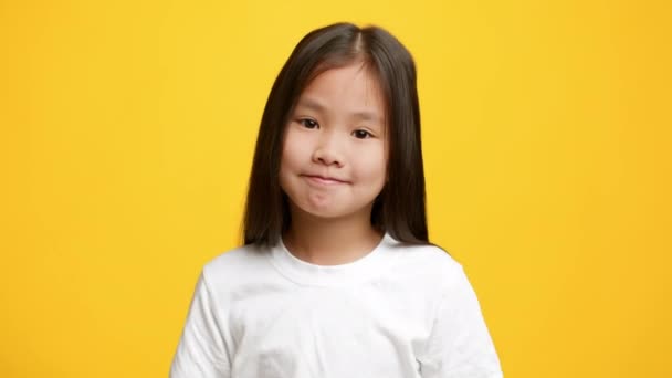 Безмозглая азиатская маленькая девочка пожимает плечами, позируя на жёлтом фоне — стоковое видео