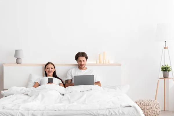 Χαρούμενη ευρωπαϊκή νεαρή γυναίκα δακτυλογράφηση στο τηλέφωνο, ο σύζυγος surfing στο διαδίκτυο στο φορητό υπολογιστή στο κρεβάτι — Φωτογραφία Αρχείου