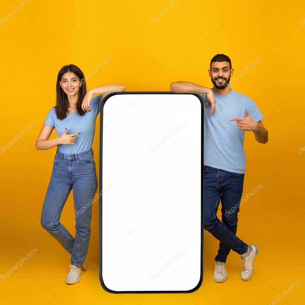 Great mobile app. Young arab couple pointing at big smartphone with empty screen mockup, promoting app or website