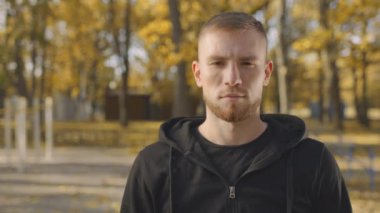 Genç, sakallı, siyah kapüşonlu bir adam sonbahar parkındaki boş alana bakıyor.