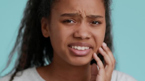 Svart tonåring flicka lider av tandvärk berörande kind, blå bakgrund — Stockvideo
