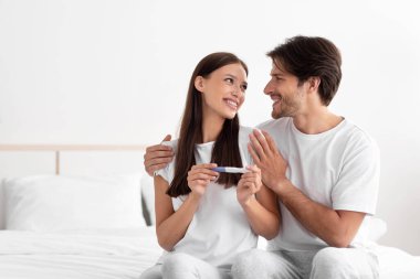 Hamilelik testi olan mutlu, heyecanlı beyaz erkek ve kadın yatak odasında neşe içinde bekliyor.