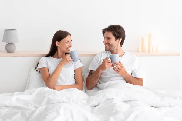 Mosolygó európai fiatal pár felébredni, és élvezze a friss kávét ül az ágyon a fehér hálószobában — Stock Fotó