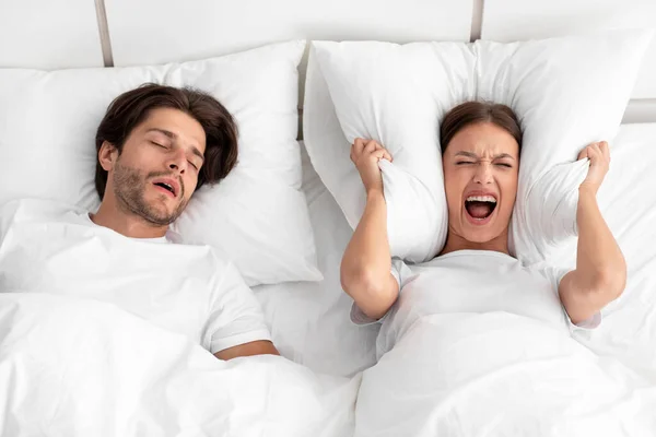 Unhappy european millennial woman awake lying in bed, shouting, covering ears with pillow — Stock Photo, Image