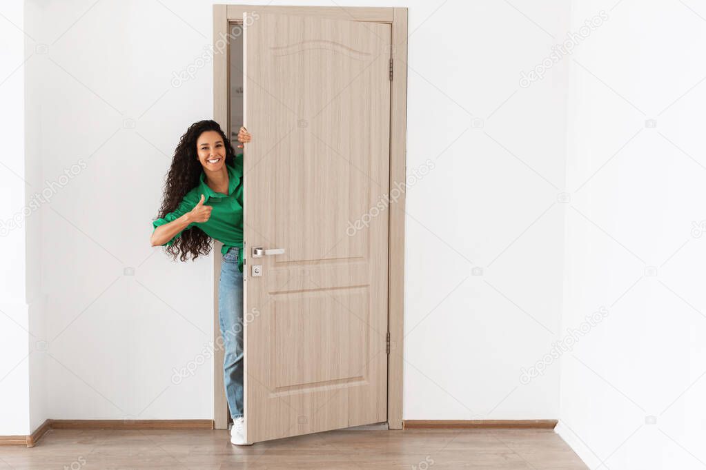 Cheerful lady looking out of door showing thumbs up gesture