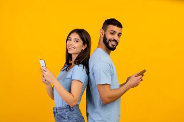 Modern yaşam tarzı. Sırt sırta duran, cep telefonlarını tutan ve kullanan pozitif Arap çift, sarı arka plan.