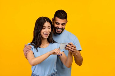 Neşeli Arap çift akıllı telefon ekranına bakıyor, yeni bir uygulama kullanıyorlar, sarı stüdyo arkaplanının başında duruyorlar.