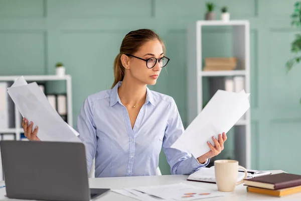 Empresária focada trabalhando em laptop e segurando relatórios, sentada no local de trabalho no interior do escritório, lendo documentos — Fotografia de Stock