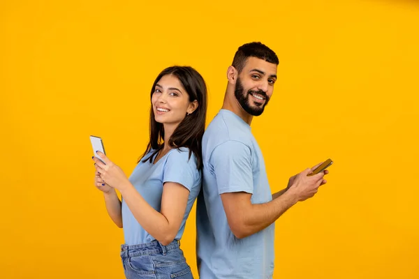 Style de vie moderne. Couple arabe positif debout dos à dos, tenant et utilisant leurs téléphones mobiles, fond jaune — Photo