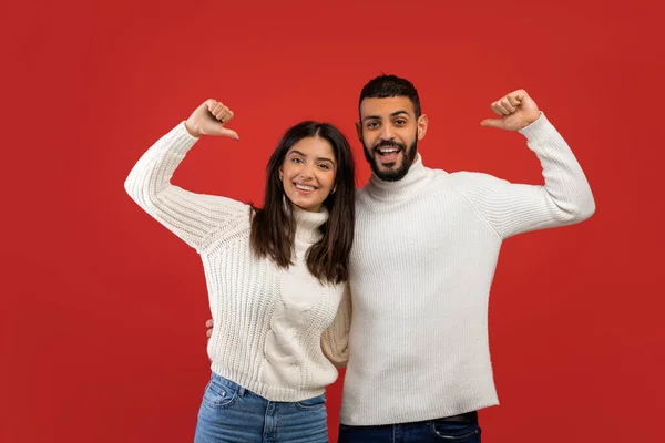 Feliz joven árabe cónyuges en suéteres blancos posando y señalando pulgares sobre sí mismos, fondo rojo —  Fotos de Stock