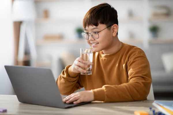 Zdrowy koreański nastolatek pijący wodę podczas korzystania z laptopa w domu — Zdjęcie stockowe