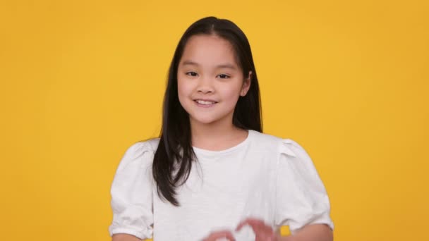 Любовь и милосердие. Милая маленькая азиатка, показывающая сердечный жест и улыбающаяся в камеру, выражающая доброту — стоковое видео