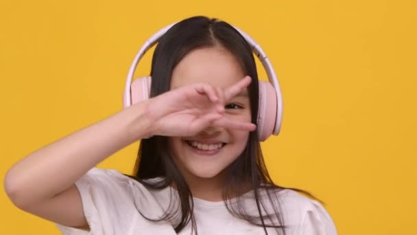 快乐的亚洲小女孩头戴耳机，听着音乐和跳舞，在她的眼前挥动签名，对着相机微笑 — 图库视频影像