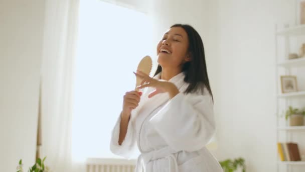 Jovem mulher asiática positiva em roupão de banho cantando em escova como microfone, desfrutando de procedimentos matinais, dançando no banheiro — Vídeo de Stock