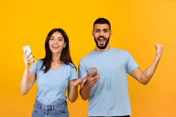 Online-Lotterie. Überglückliche junge arabische Männer und Frauen jubeln mit ihren Smartphones über den Erfolg, gelber Hintergrund — Stockfoto