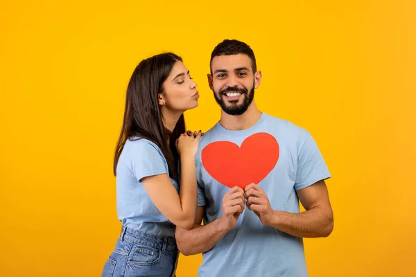 День Святого Валентина концепция. Портрет любящей арабской пары, держащей карточку из красной бумаги, молодой женщины, целующей своего мужа — стоковое фото