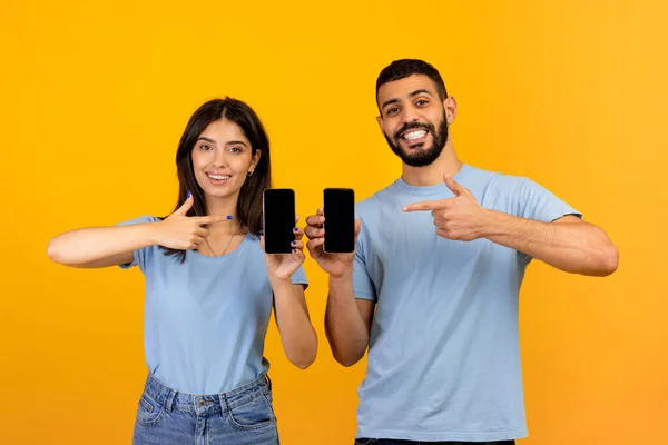 Нам нравится это приложение. Счастливые арабские супруги держат и представляют смартфоны с черными экранами, указывая на гаджеты — стоковое фото