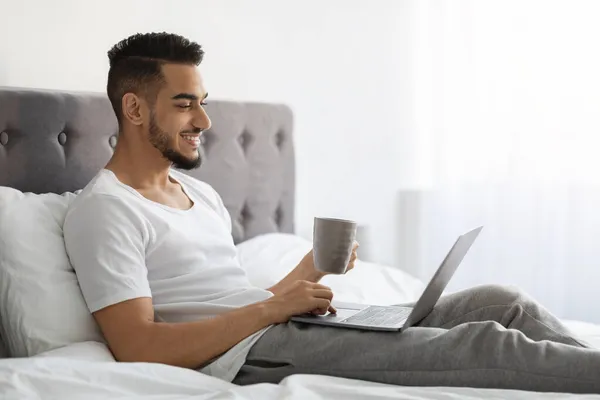 Szczęśliwy Młody Bliski Wschód Mężczyzna relaks w łóżku z laptopem i kawy — Zdjęcie stockowe