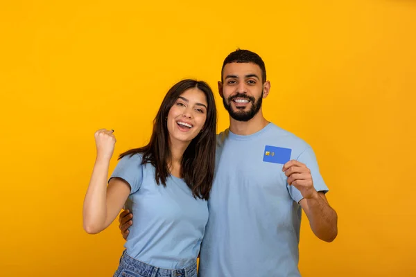 Pareja árabe muy alegre con tarjeta de crédito haciendo gesto SI, promoviendo compras sin contacto sobre fondo amarillo —  Fotos de Stock