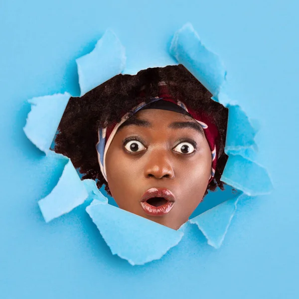 Incroyable choqué jeune femelle noire avec la bouche ouverte et les yeux, regarde à travers le trou dans le papier bleu — Photo