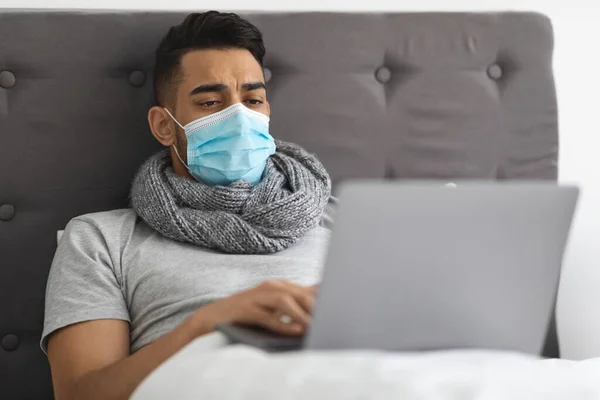 Больной молодой араб в медицинской маске работает с ноутбуком в постели — стоковое фото