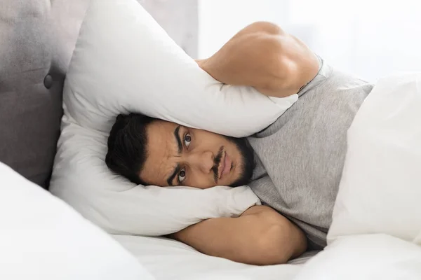 Podrážděný mladý arabský muž ležící v posteli a zakrývající hlavu polštářem — Stock fotografie