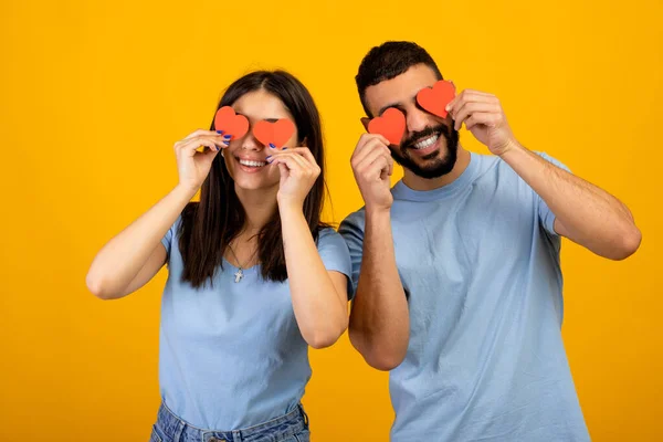 Aveuglé par l'amour. Jeune couple arabe romantique tenant des coeurs en papier rouge couvrant les yeux, posant sur fond jaune — Photo