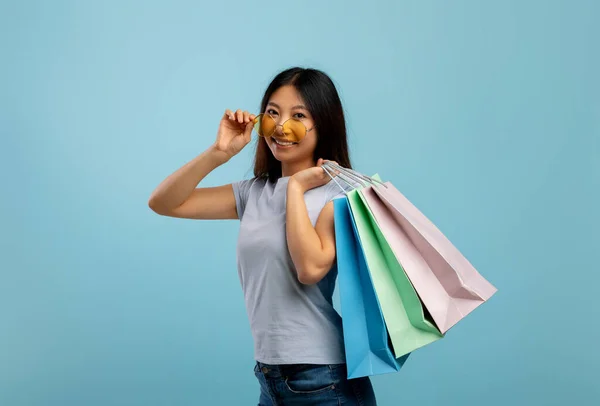 Speciale aanbieding. Jonge Aziatische dame aanraken zonnebril met kleurrijke boodschappentassen met aankopen, blauwe achtergrond — Stockfoto