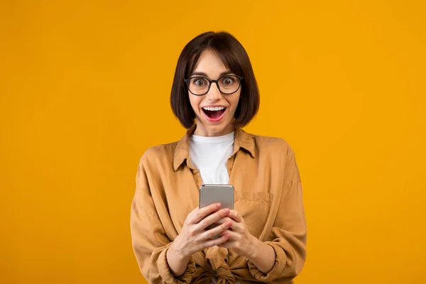 Omg, increíble. Mujer sorprendida sosteniendo y usando smartphone, recibiendo messag, fondo amarillo del estudio — Foto de Stock