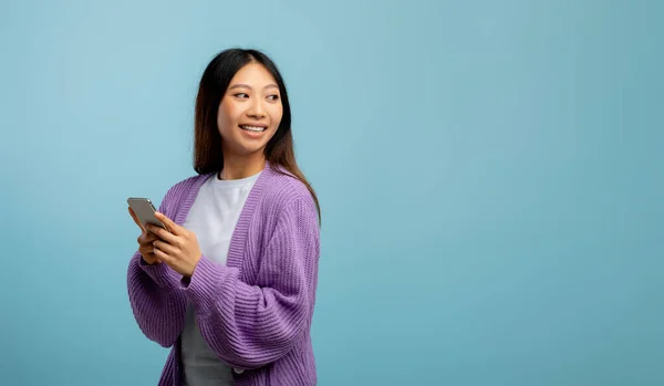 Fröhliche asiatische Dame mit neuestem Handy blickt zurück auf Kopierraum über blauem Studiohintergrund, Panorama — Stockfoto
