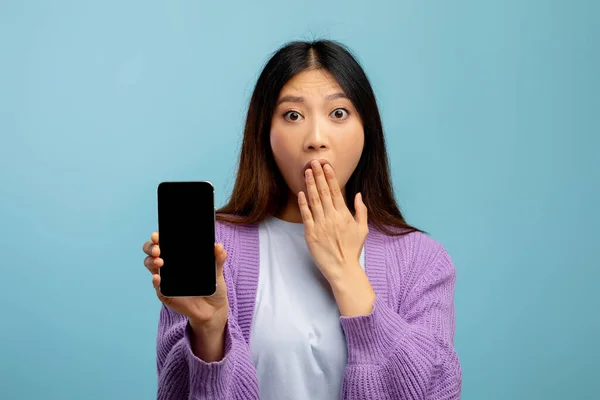 Geschokt aziatische dame tonen smartphone met leeg scherm en het bedekken van haar mond, staan op blauwe achtergrond, mockup — Stockfoto