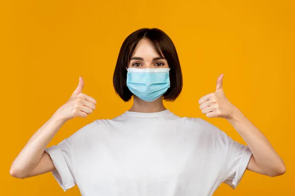 Носить маску концепции. Портрет молодой женщины в медицинской маске и жесты большие пальцы вверх, желтый фон — стоковое фото