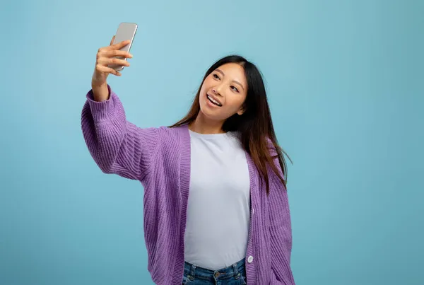 Schattig aziatisch dame nemen selfie op smartphone, het hebben van speelse stemming en glimlachen op webcamera, blauwe achtergrond — Stockfoto