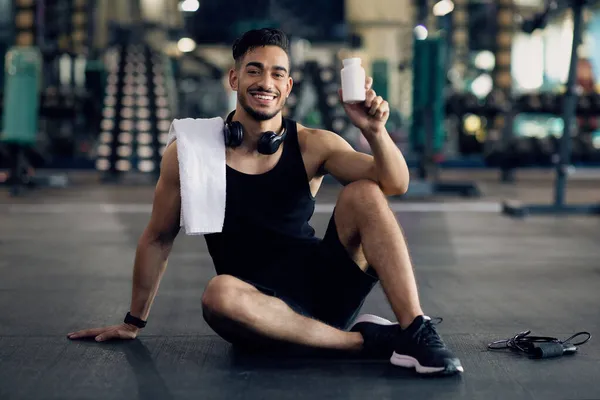Homem árabe muscular jovem segurando recipiente com pílulas de suplemento, posando no ginásio — Fotografia de Stock