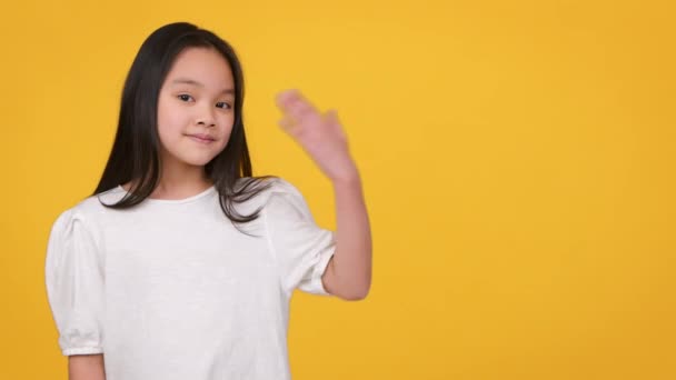 Bedårande liten asiatisk flicka sträcker ut ena handen åt sidan, visar något över orange studio bakgrund — Stockvideo