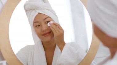 Kafasında havluyla bornoz takan genç, güzel Koreli kadın yüzünü losyonla temizliyor, yansımasına gülümsüyor.