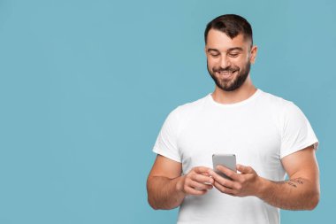 Neşeli olgun beyaz adam mavi arka planda izole akıllı telefondan mesaj yazıyor.