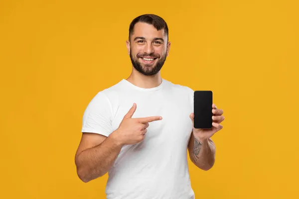 Lachend zelfverzekerd knappe volwassen europese man blogger in wit t-shirt met vinger op smartphone met leeg scherm — Stockfoto