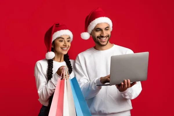Onlineförsäljning. Arabiska par bär Santa Hattar håller laptop och shoppingväskor — Stockfoto