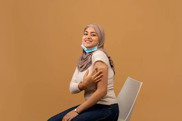 Covid-19ワクチン接種キャンペーン。コロナウイルスワクチン注射後の腕を示すヒジャーブのイスラム教徒の女性,茶色の背景 — ストック写真