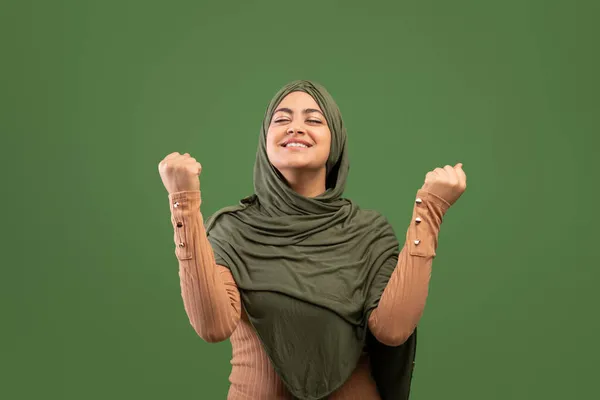 ユーフォリックアラブ女性でhihab祝う成功とともに手を上げ、拳を握りしめ,濃い緑の背景 — ストック写真